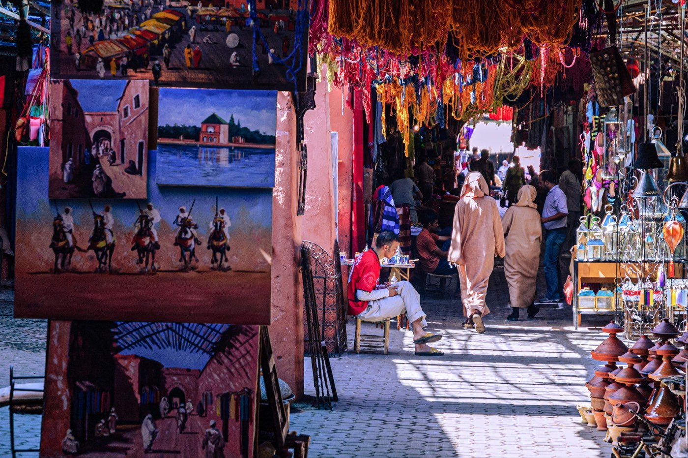 A souk lane hung with dyed wool and lanterns, paintings stacked against the wall and shoppers passing in the dappled light