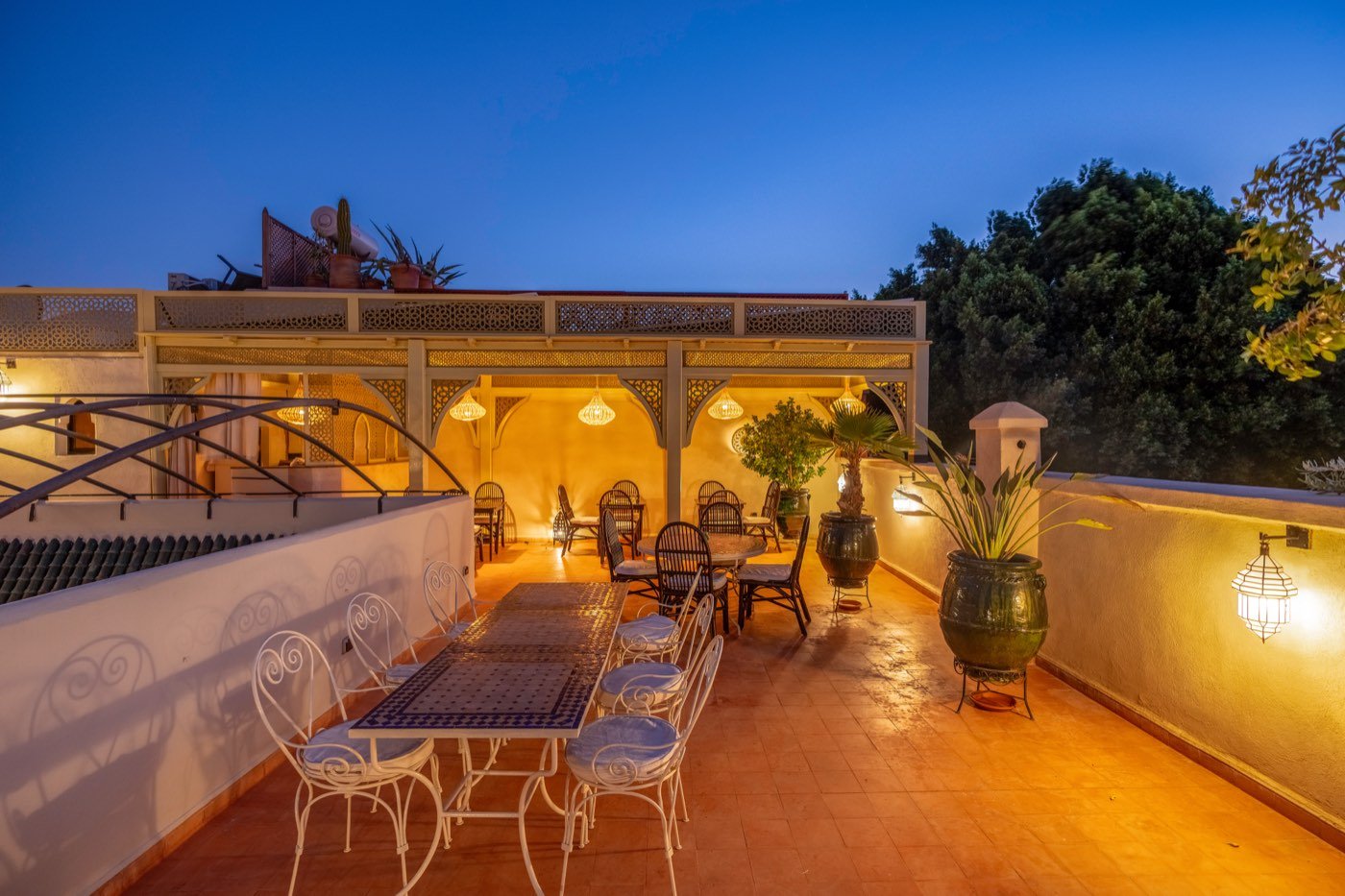 The rooftop pergola at dusk, its fretwork panels and carved brackets lit from within, a mosaic table laid beneath