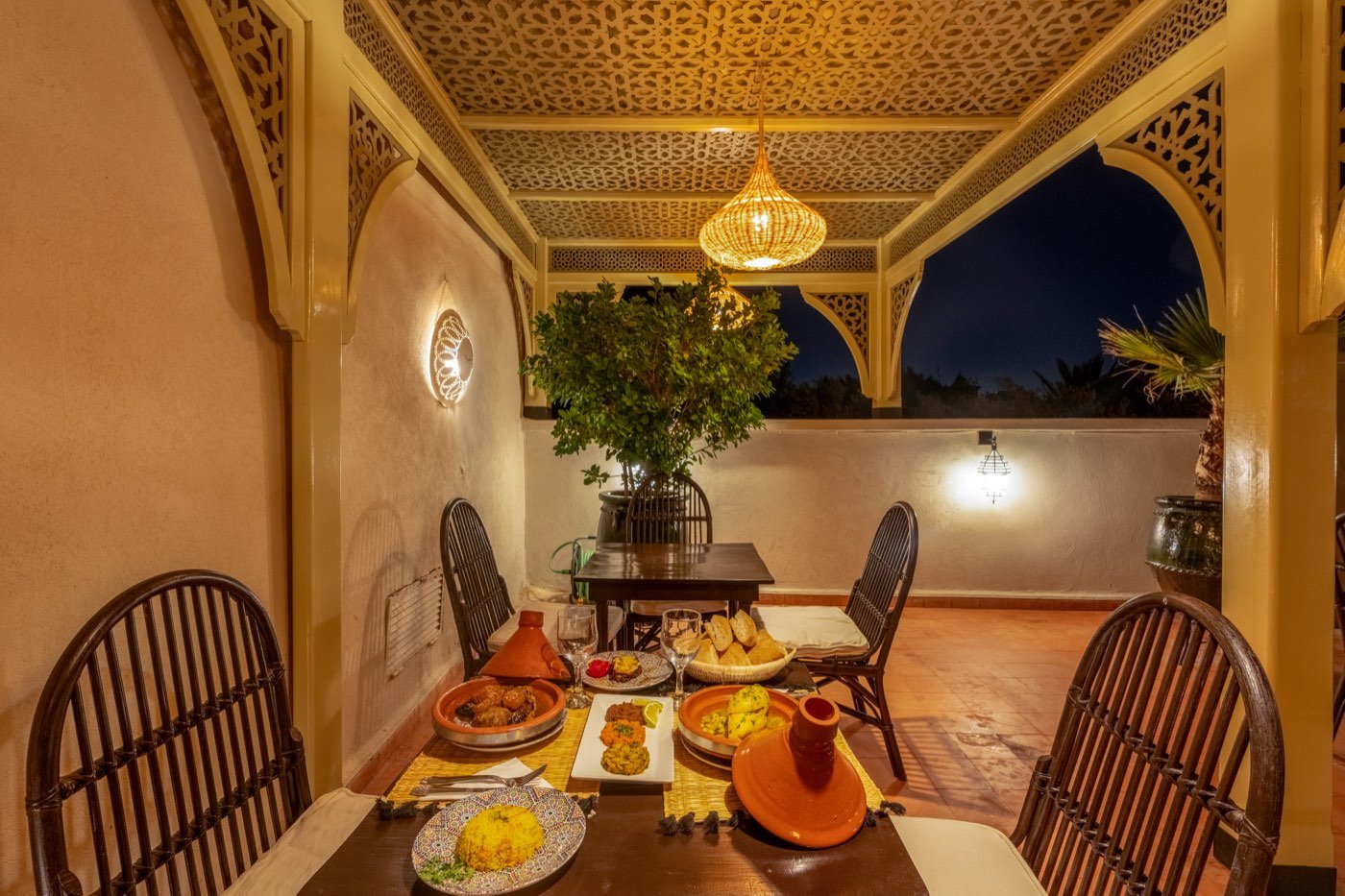 A table laid for dinner under the terrace's carved ceiling — tajines, bread and saffron rice below a woven lantern, the night sky through the arch beyond